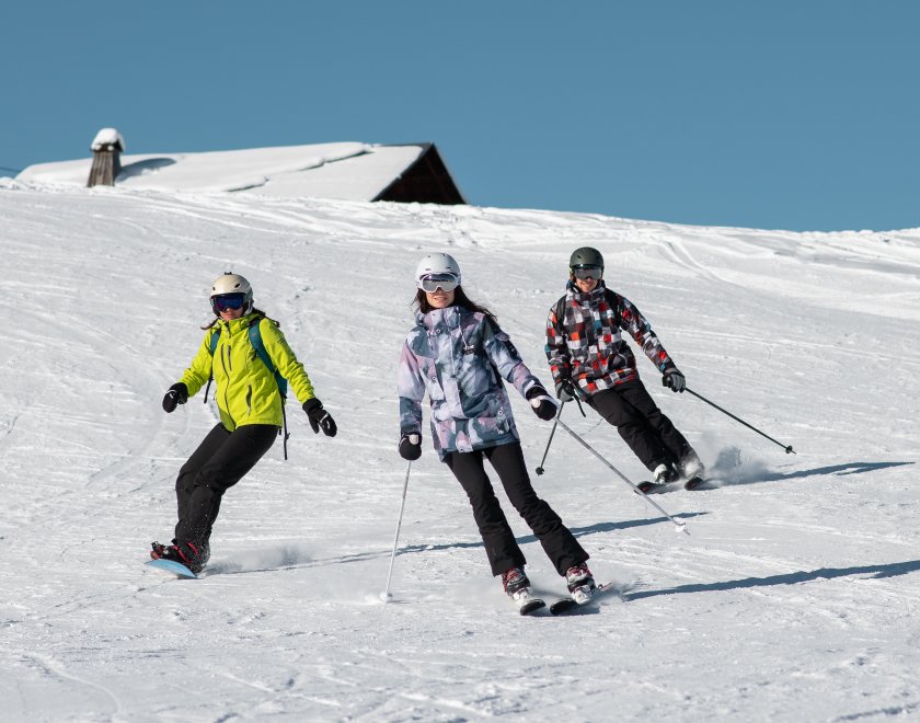 Un groupe de 3 amis déscend une piste avec un chalet dont le toît est enneigé en fond
