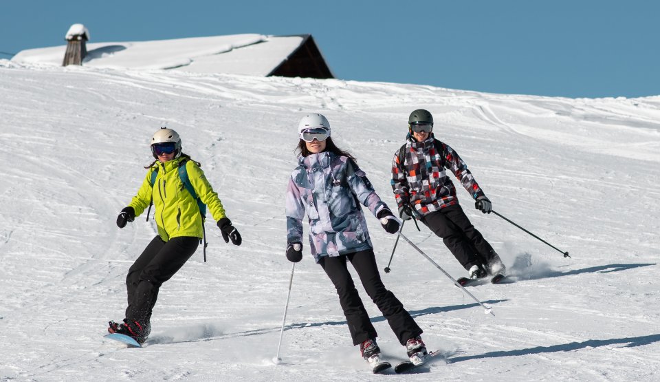 Un groupe de 3 amis déscend une piste avec un chalet dont le toît est enneigé en fond